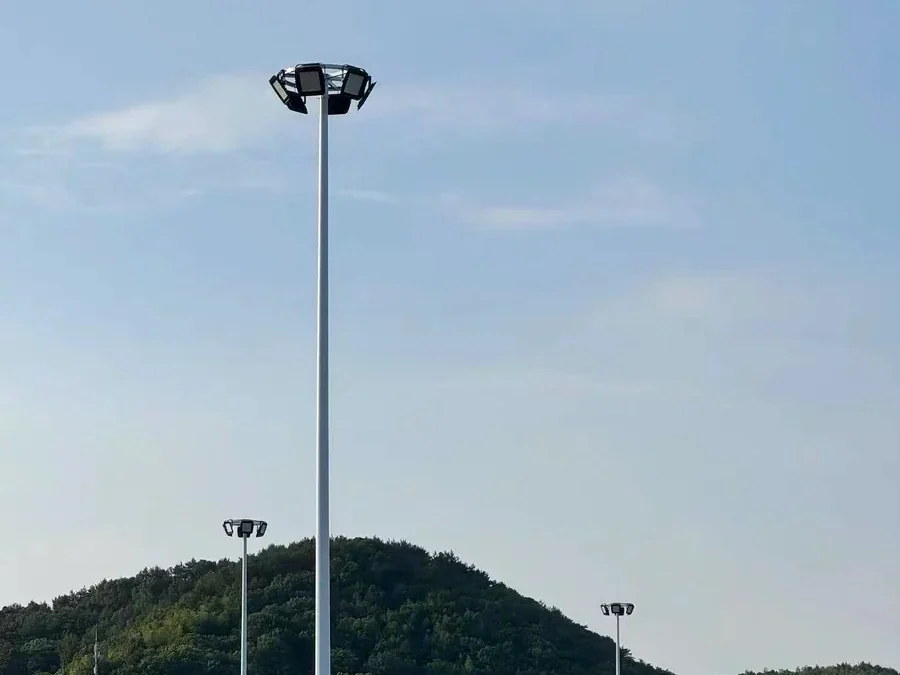 High mast lighting tower on an open yard with a wooded hillside behind, floodlights mounted in a ring at the top of the mast
