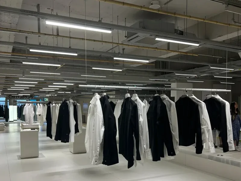 Retail floor with clothing rails lit by continuous rows of linear LED fixtures across an exposed-services ceiling