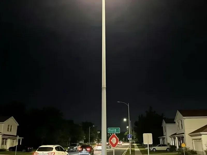 Suburban street junction at night lit by a high mast lighting tower, with a parked car and road markings visible under the light
