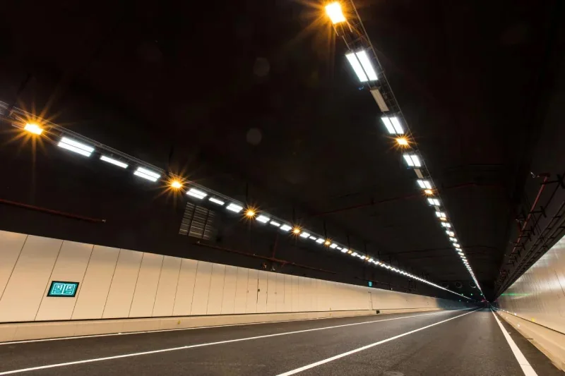Long road tunnel interior with linear LED luminaires mounted in a continuous line down the ceiling, giving even light along the driving lane